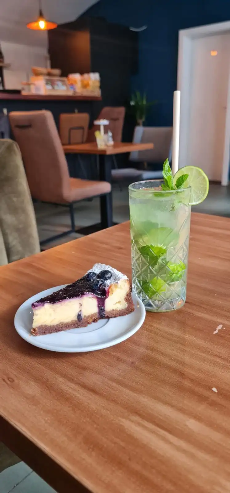 Slice of blueberry cheesecake on a white plate next to a Mojito.