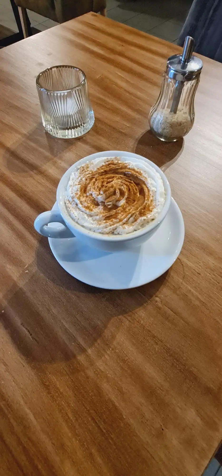 Cappuccino dusted with cocoa in a white cup on a wooden table, sugar dispenser and a small glass beside it, seen from above at an angle.