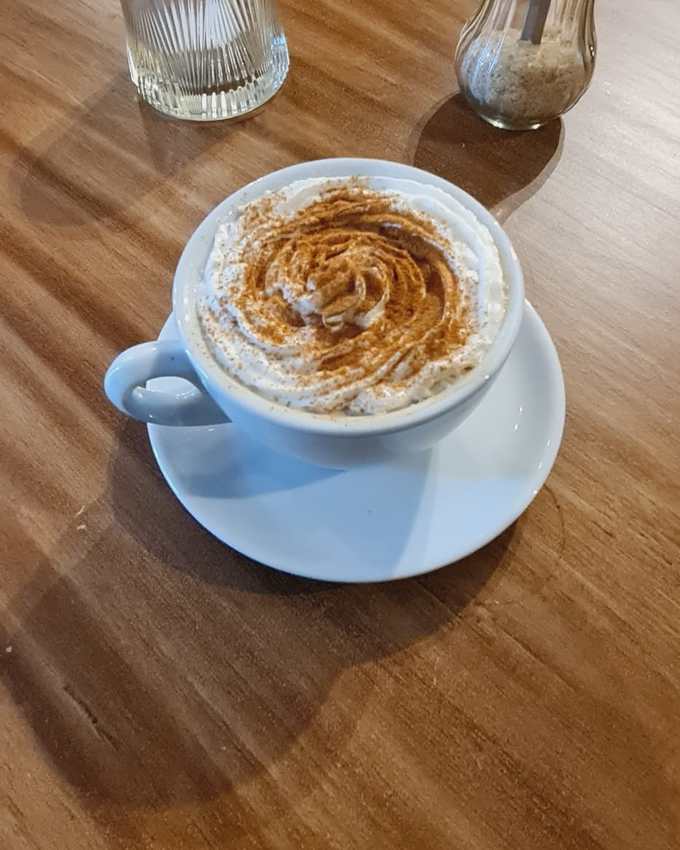 Cappuccino dusted with cocoa in a white cup on a wooden table, sugar dispenser and a small glass beside it, seen from above at an angle.