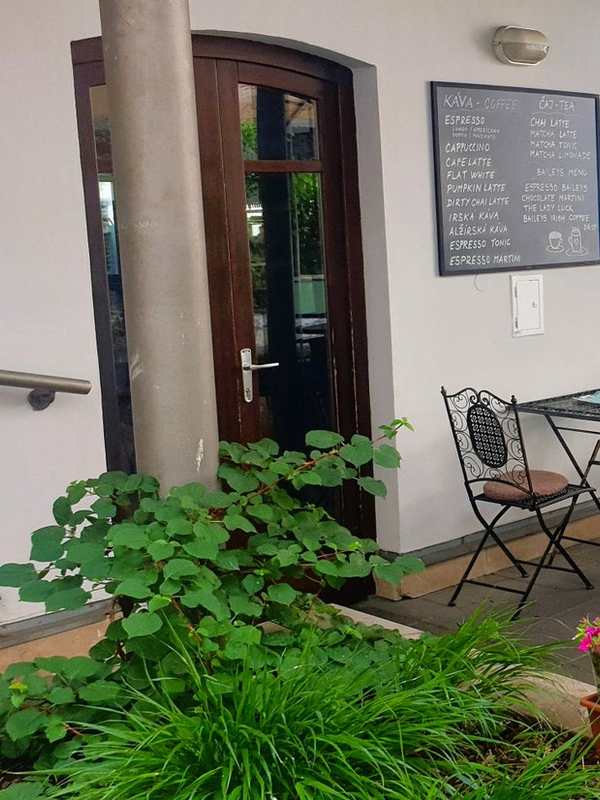 Arcade side of the courtyard: columns, bistro tables and chairs, flower boxes, arched doors of the cafe, a chalkboard on the wall.