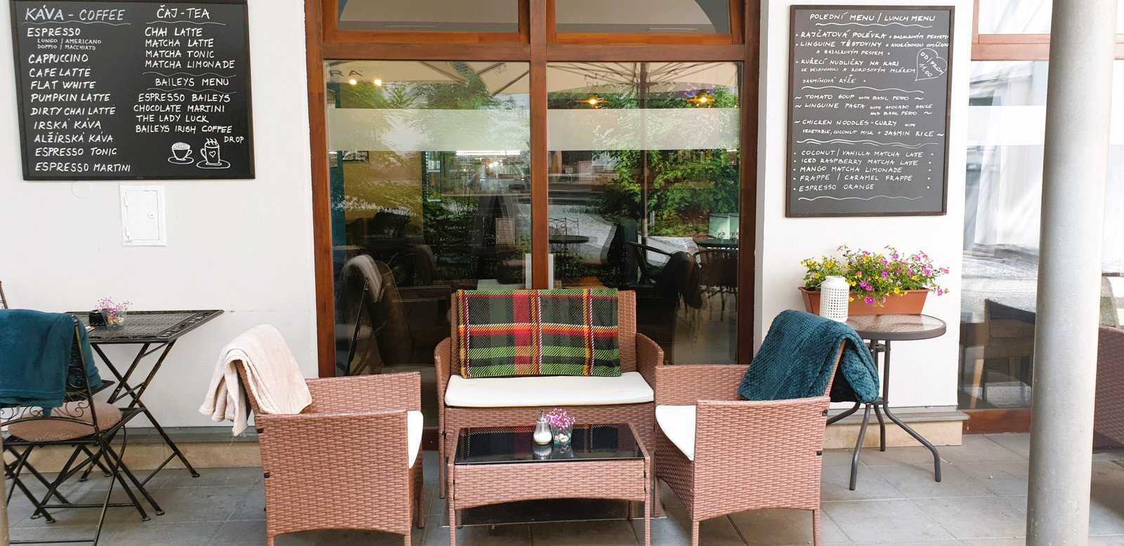 Courtyard lounge corner: rattan sofa and armchairs with plaid cushions, small table with candle, flower box, glass facade of the cafe, two chalkboard menus (KÁVA - COFFEE, ČAJ - TEA) on the wall.