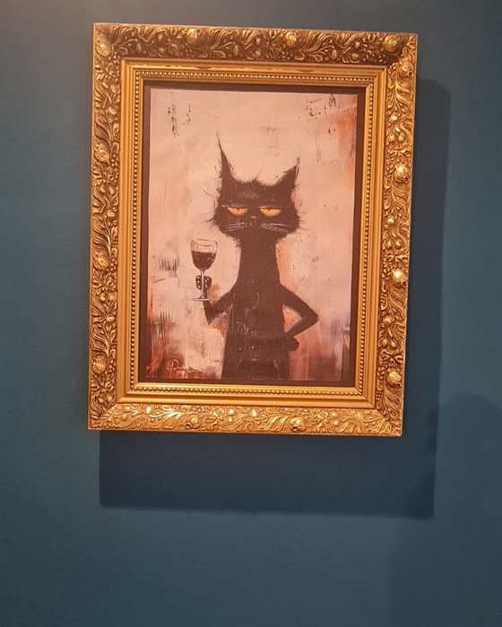 Ornate gold frame with a painting of a black cat holding a wine glass, on a dark teal wall.