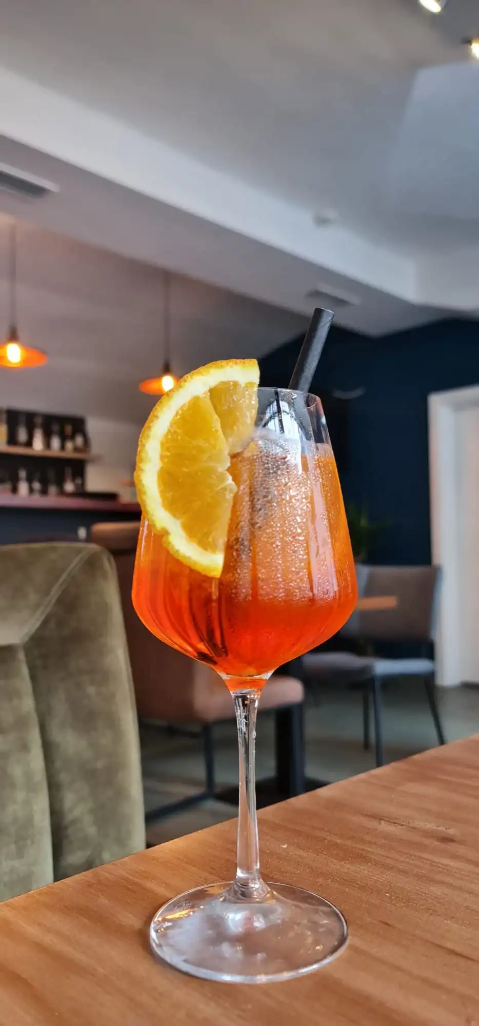 Aperol Spritz on a table with the bar, bottle shelf and pendant lamps softly blurred behind.