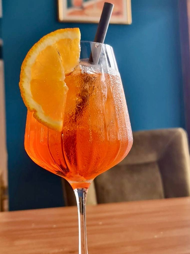 Aperol Spritz with an orange slice on a wooden table, blue wall with a framed Aperol poster and a velvet chair behind.