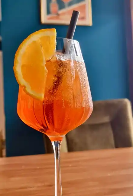 Aperol Spritz with an orange slice on a wooden table, blue wall with a framed Aperol poster and a velvet chair behind.