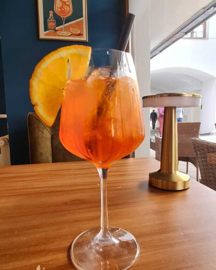 Aperol Spritz by the window, gold cordless table lamp, blue wall with Aperol poster.