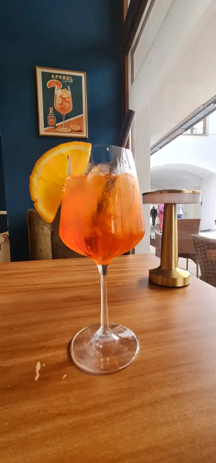 Aperol Spritz by the window, gold cordless table lamp, blue wall with Aperol poster.