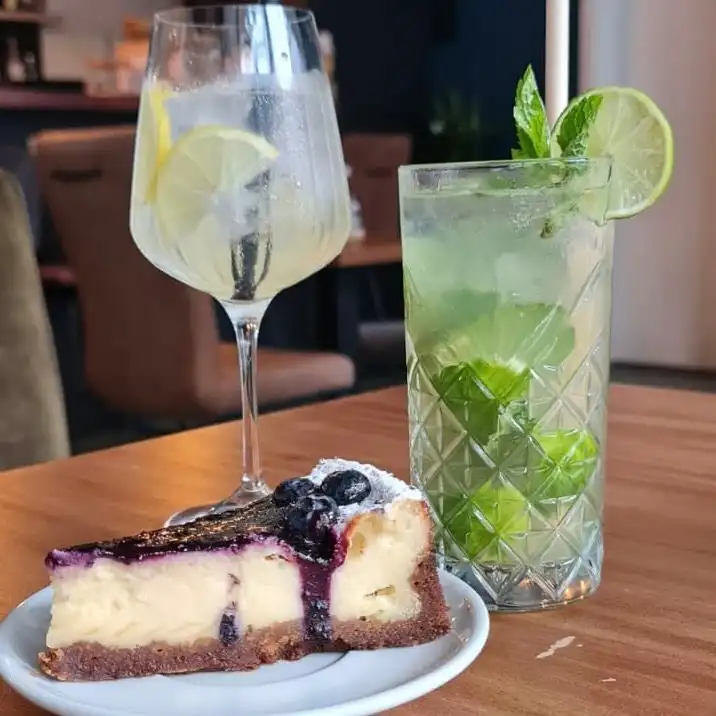 Square close crop: blueberry cheesecake in front, gin and tonic and Mojito behind, cafe interior blurred.