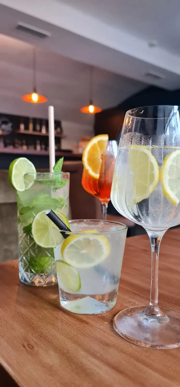 Three drinks together: Mojito, Aperol Spritz, gin and tonic in a wine glass, plus a tumbler gin and tonic, bar behind.