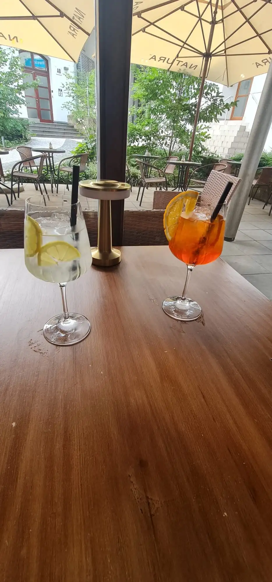 Gin and tonic and Aperol Spritz on a table by the window, courtyard with parasols and greenery outside.