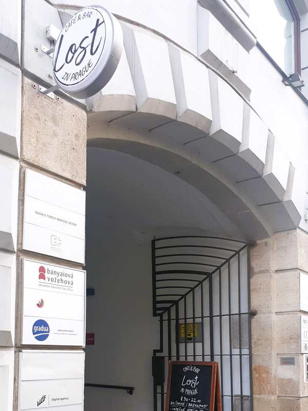 Street entrance on Lazarská: stone archway with a wrought-iron gate, round "Lost in Prague" sign above, company plaques, chalkboard A-board inside, cobblestones. A blue arrow is drawn on the photo pointing to the door.