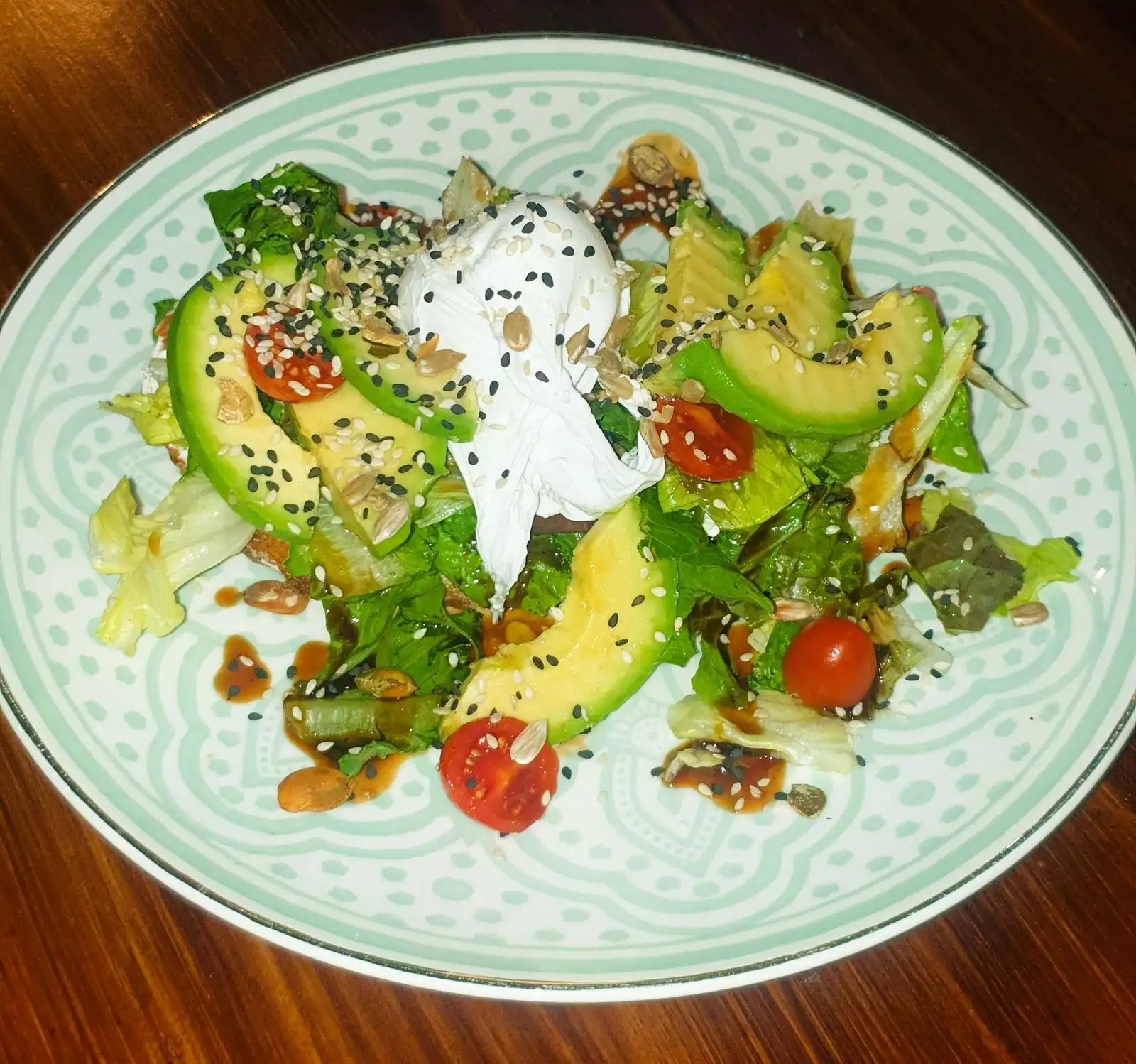 Avocado bread top-down: avocado slices, poached egg, salad, cherry tomatoes, seeds on a mint patterned plate.