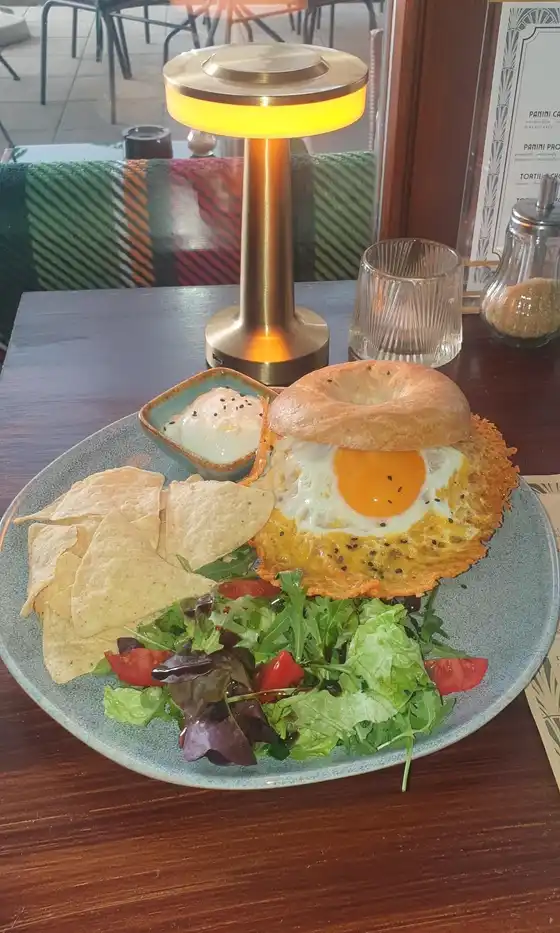 BEEF BAGEL: bagel with pulled beef and a cheddar-crusted fried egg, nachos, salad and a dip on a teal plate; gold table lamp and the courtyard behind.