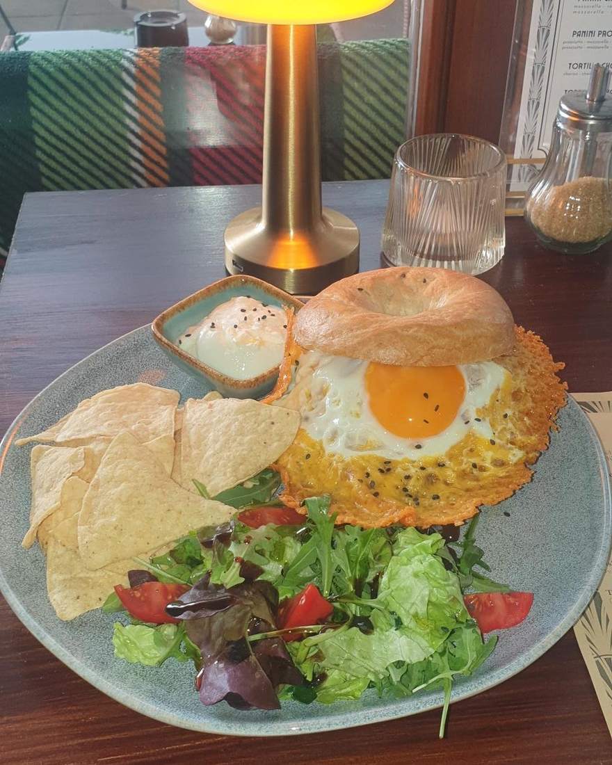 BEEF BAGEL: bagel with pulled beef and a cheddar-crusted fried egg, nachos, salad and a dip on a teal plate; gold table lamp and the courtyard behind.
