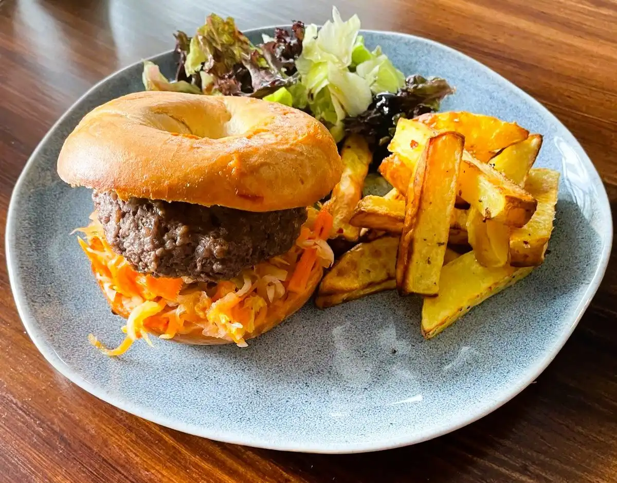 Beef burger in a toasted bagel with kimchi and carrot, roasted potato wedges and salad on a blue plate.