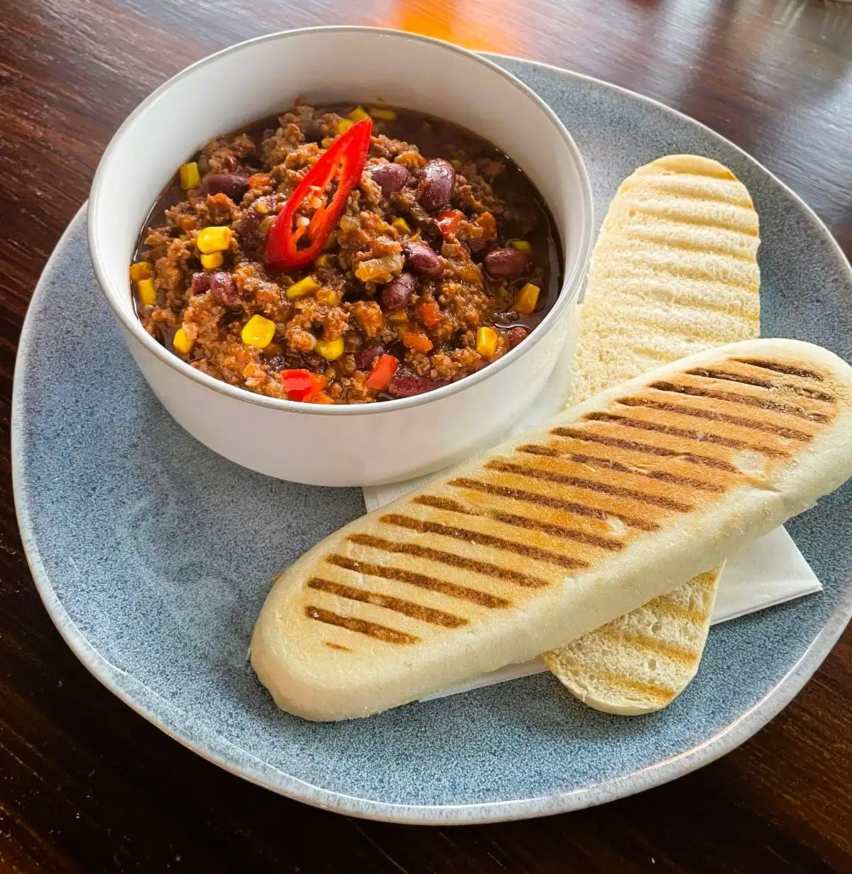Chilli con carne in a white bowl with a red chilli on top and two grilled panini toasts on a blue stoneware plate.