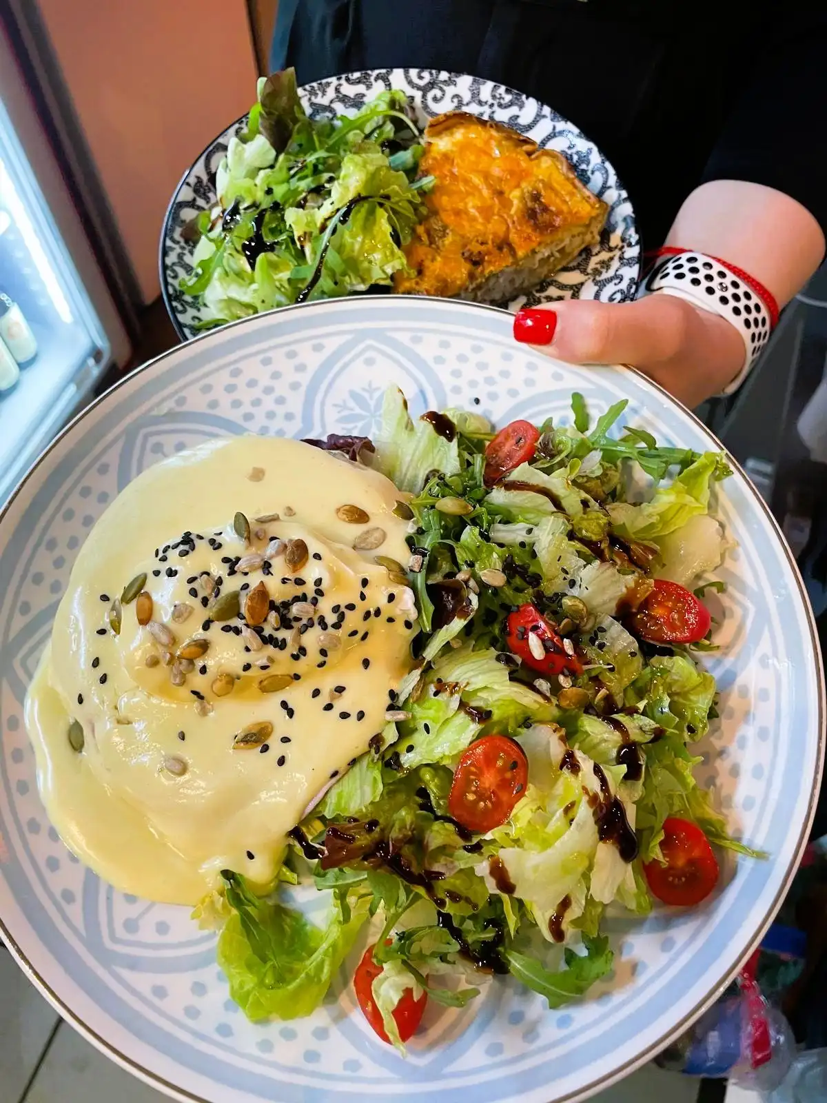 Egg Benedict on a bagel with hollandaise, black sesame, salad and cherry tomatoes on a patterned plate held by a hand with red nails; a quiche plate behind.