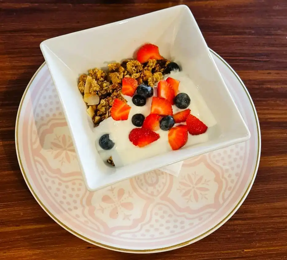 House-made granola with yoghurt, strawberries and blueberries in a square white bowl on a patterned plate.