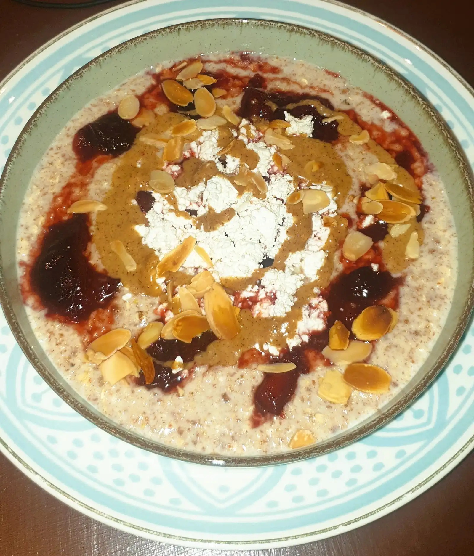 Oatmeal with stewed plums, nut butter, curd and almond flakes in a round bowl, top-down.