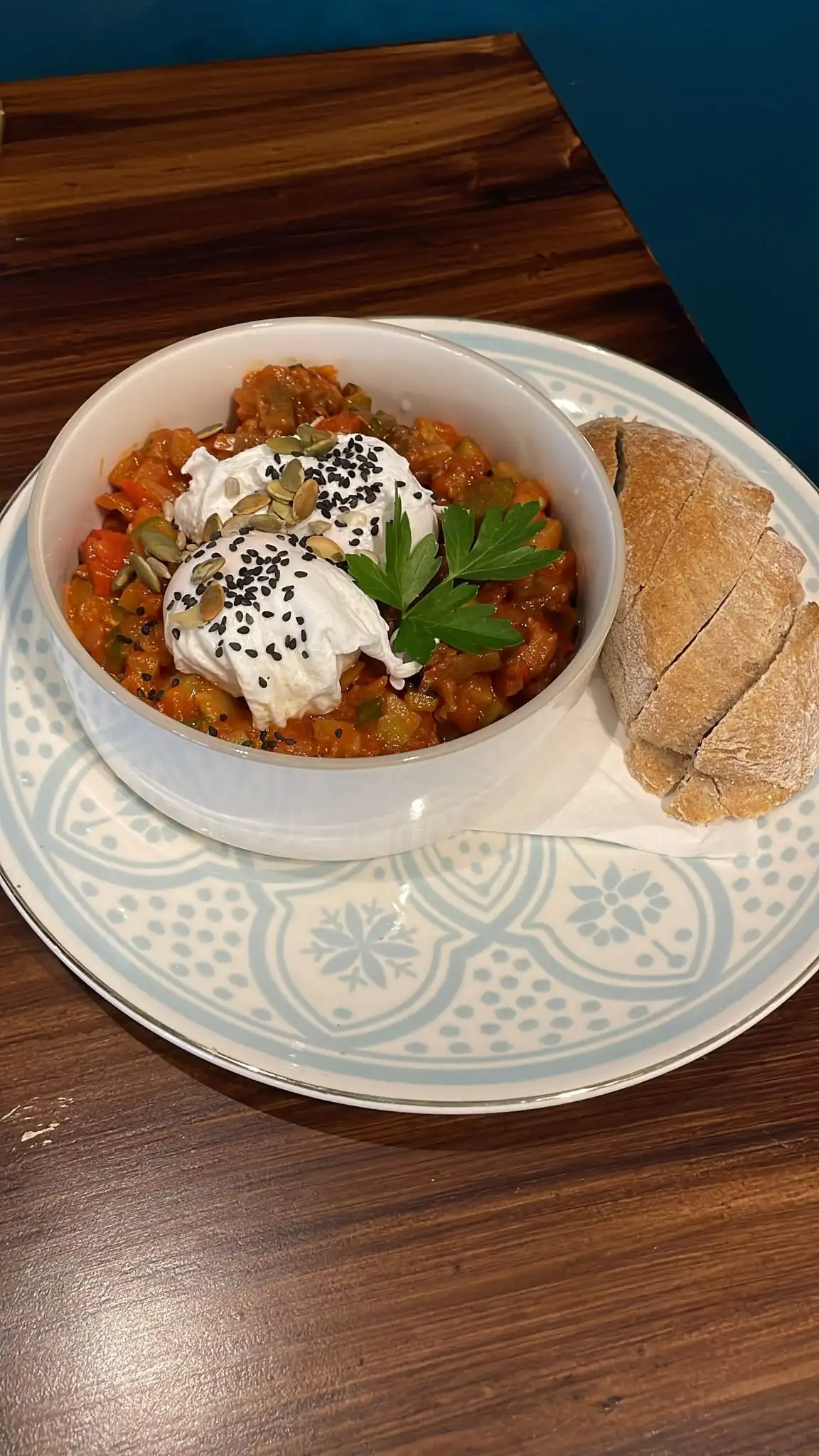 Ratatouille in a white bowl topped with poached eggs, yoghurt, black sesame and parsley, sourdough slices on the side.