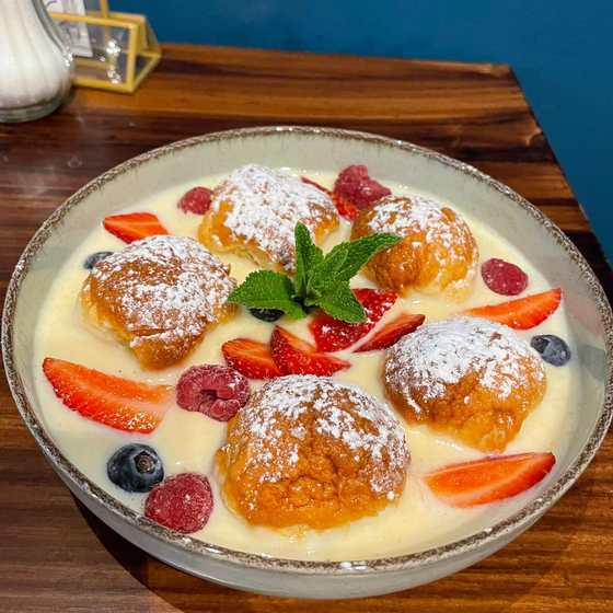 Sweet buns dusted with sugar in vanilla custard with strawberries, raspberries, blueberries and mint, top-down on a dark wooden table.