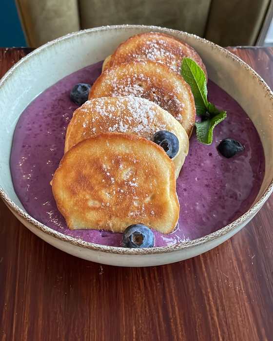 Four yeast pancakes dusted with sugar on purple blueberry-mascarpone cream with blueberries and mint, in a stoneware bowl.