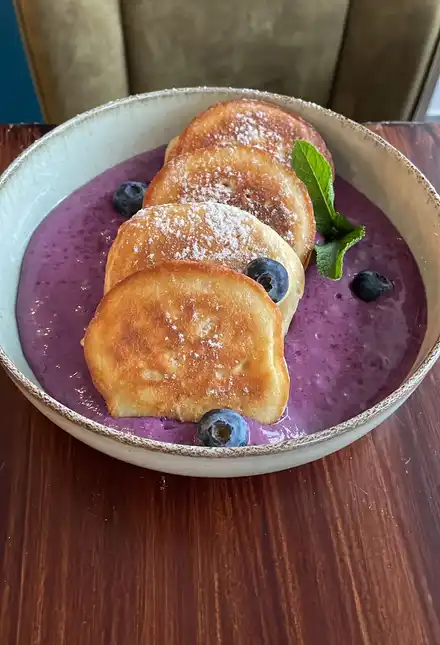 Four yeast pancakes dusted with sugar on purple blueberry-mascarpone cream with blueberries and mint, in a stoneware bowl.