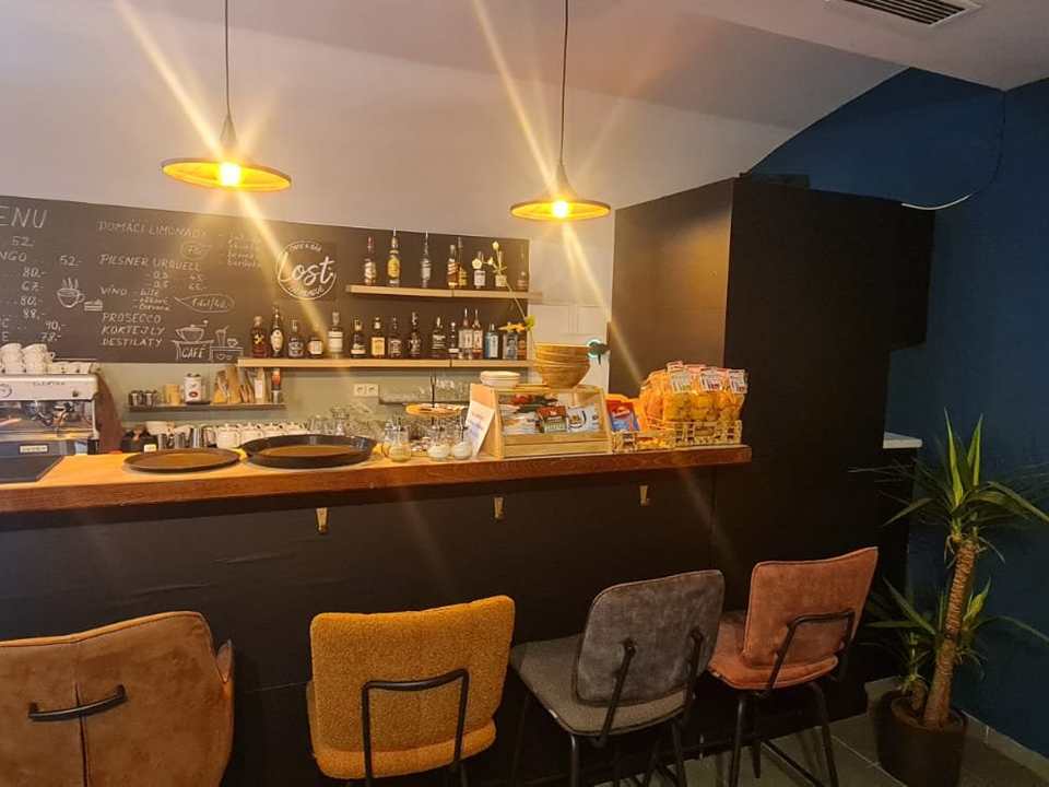 Bar front, 2024: long counter with coloured chairs, espresso machine, chalkboard menu wall, bottle shelf, pendant lamps, plant.